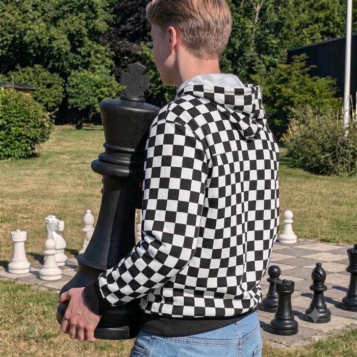 Dreng i sort og hvid skakternet hoodie, der står udendørs med en stor, sort skakbrik i hænderne.   