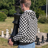 Dreng i sort og hvid skakternet hoodie, der står udendørs med en stor, sort skakbrik i hænderne.   