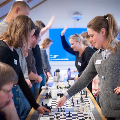 Et fotografi af en gruppe voksne, der spiller skak ved lange borde i et lyst lokale. En kvinde i forgrunden peger på en brik på et af skakbrætterne, mens flere personer i baggrunden rækker en hånd i vejret. Stemningen virker engageret og lærerig.